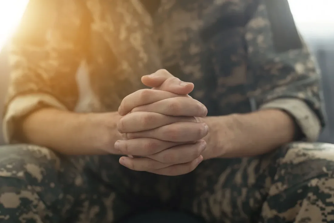 A service member sits with hands clasped in warm early light